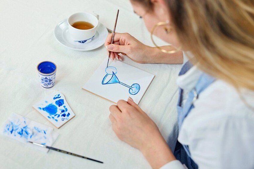 A person painting a blue and white design on paper, with a cup of coffee and paint containers on the table. 2