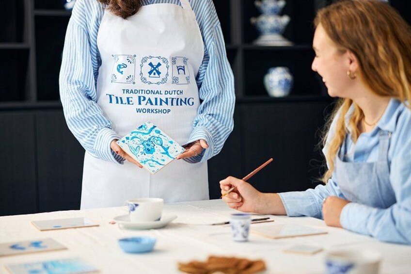 A person in a striped shirt and apron holding a tile painting, standing at a table with a woman painting tiles. 3