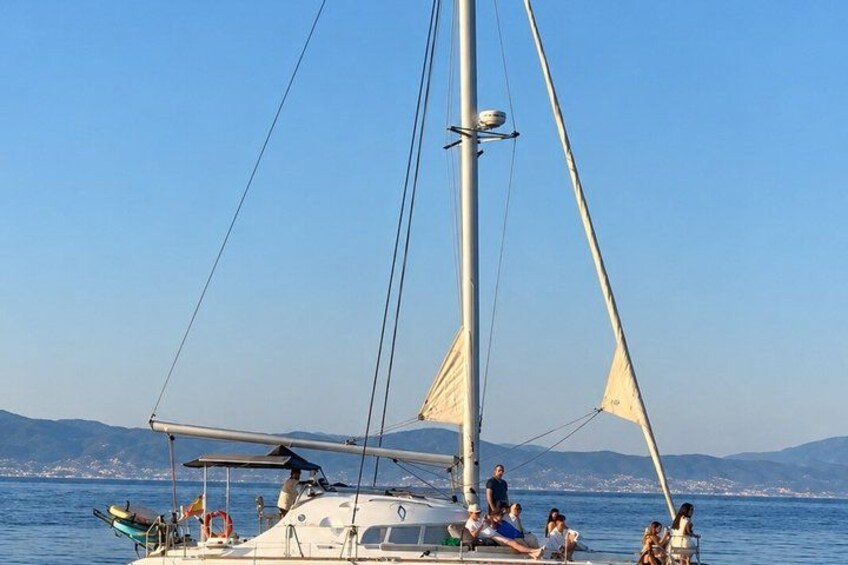 A sailboat with people on board, anchored near a coastline with mountains in the background. 2