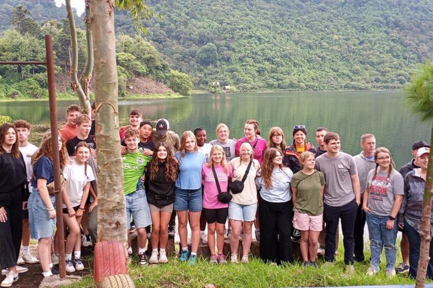 A group of people standing by a lake with trees and hills in the background. 5