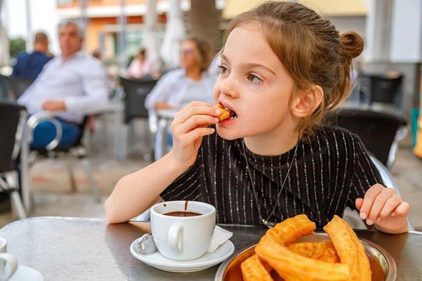 A person sitting at an outdoor cafe table with a cup of coffee and a plate of fritters. 3