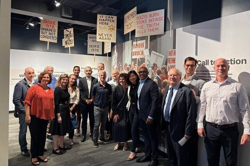 A group of people standing in an indoor exhibition space with informational signs and banners. 3