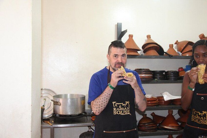 Two people wearing aprons with the text 'KABAB SOUSS' in a kitchen setting with pots and pans on the counter. 2