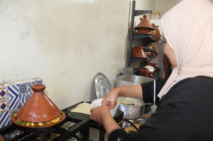 A person cooking with a traditional clay pot on a stove, surrounded by shelves with various kitchen utensils and cookware. 3