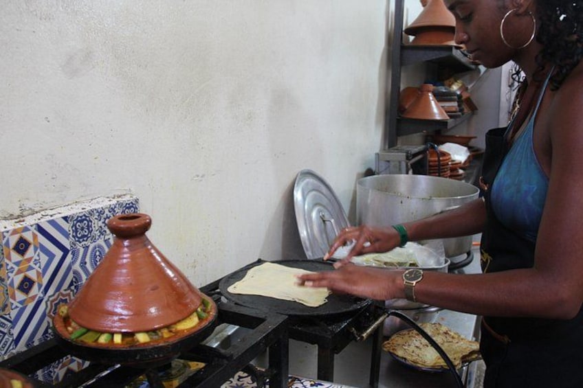 A person cooking with a flatbread on a portable stove, a clay pot, and a tiled wall with decorative patterns. 5