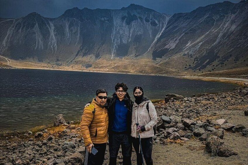 Three people standing on a rocky shore with a lake and mountains in the background. 4
