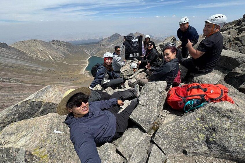 Un grupo de personas descansando en la cima rocosa de una montaña, con cielo despejado y montañas distantes al fondo. 1