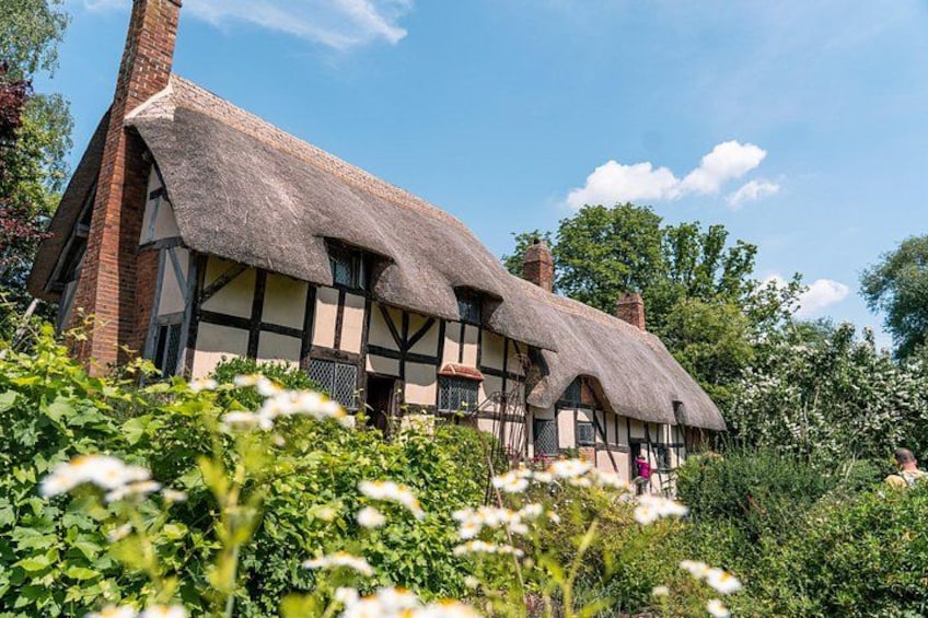 A traditional half-timbered house with a thatched roof, surrounded by lush greenery and flowering plants. 1