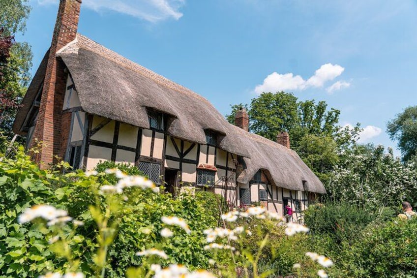 A traditional thatched-roof house with a chimney, surrounded by lush greenery and flowering plants. 1