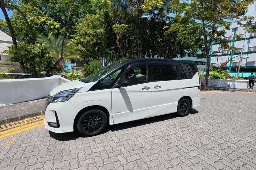 A white minivan parked on a paved area with trees and buildings in the background. 2