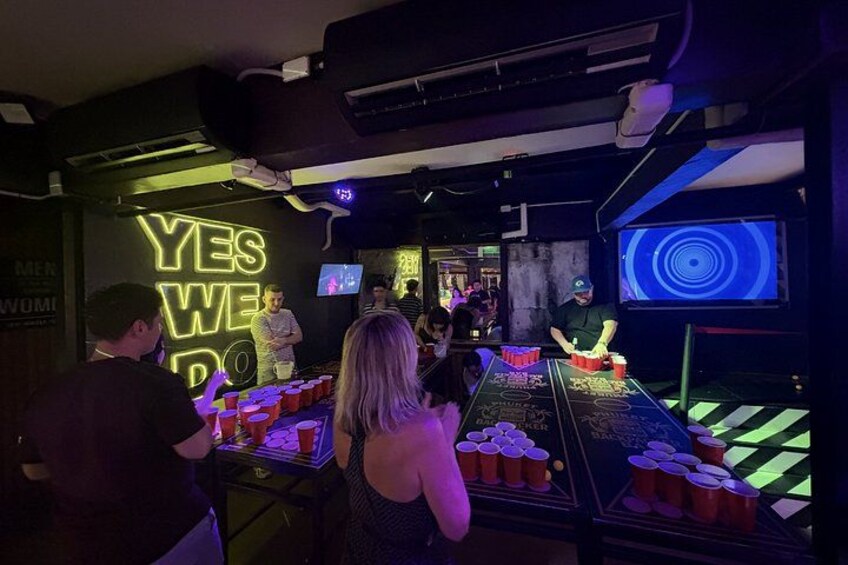 A bar with a neon sign, a pool table, and people playing beer pong. 2