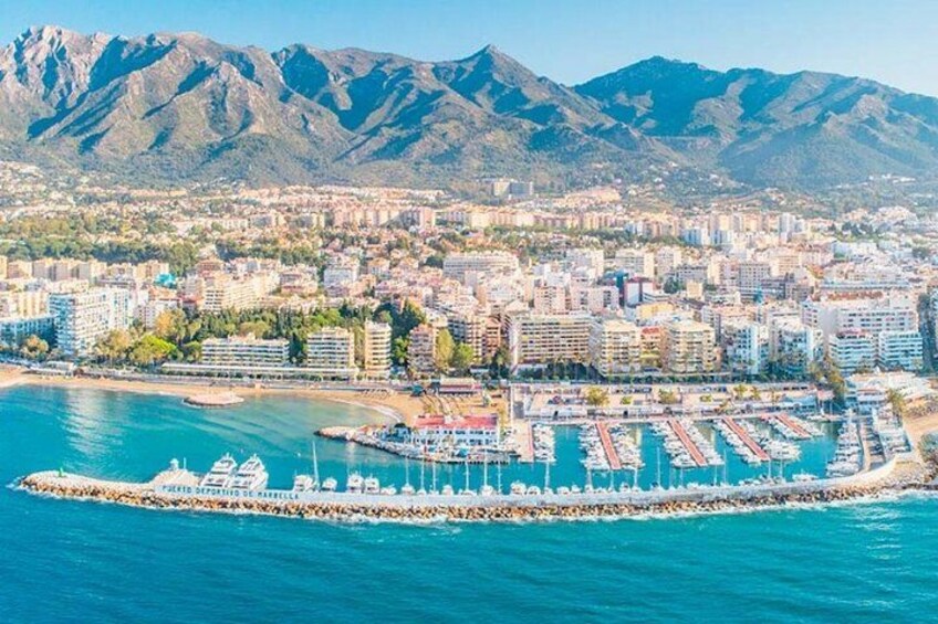 A coastal city with a marina, buildings, and mountains in the background. 1