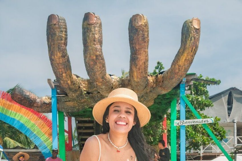 Une personne portant un chapeau de paille et un collier, debout devant une grande sculpture d’arbre à plusieurs troncs. 3