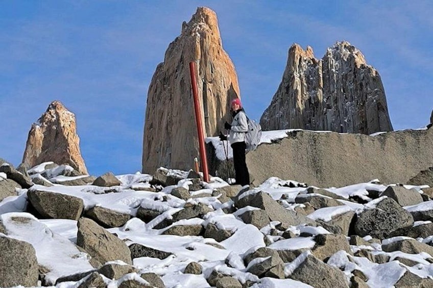 A hiker on a rocky mountain with snow patches, a red pole, and two prominent peaks in the background. 3