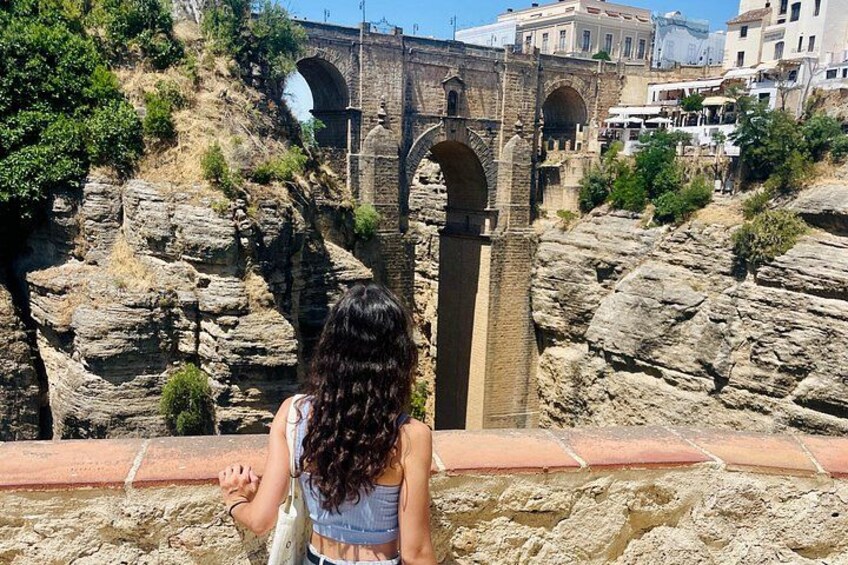 A person overlooking a stone bridge with arches, rocky terrain, and buildings in the background. 2