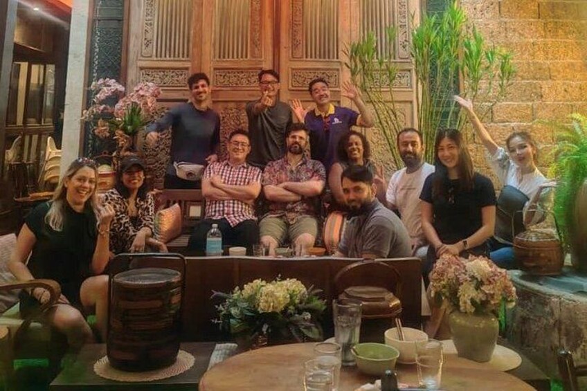 A group of people seated at a table with a rustic interior, surrounded by plants and decorative items. 2