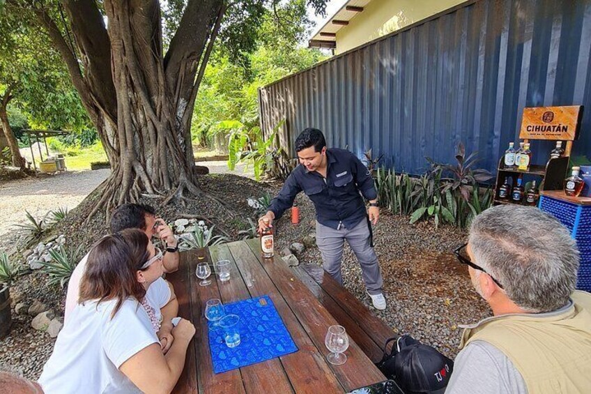 A group of people sitting at a wooden table outdoors, a man standing with a bottle, and a sign that reads 'Cihuatán'. 2