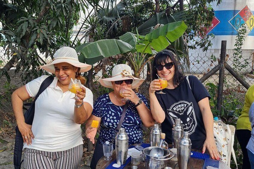 Three people standing outdoors with drinks, a table with bottles and glasses, and a fence with greenery in the background. 3