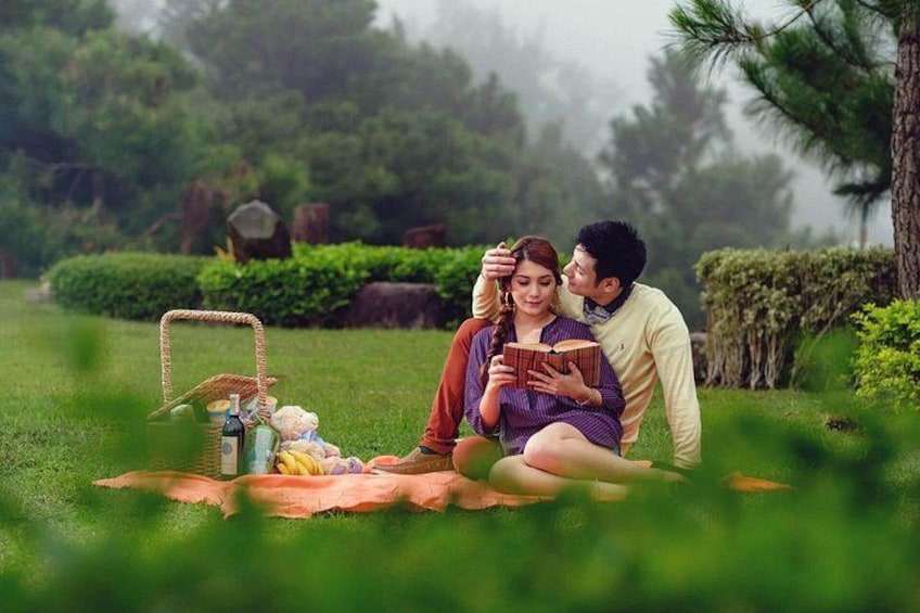 A couple sitting on a blanket in a park with a picnic basket and food items. 1