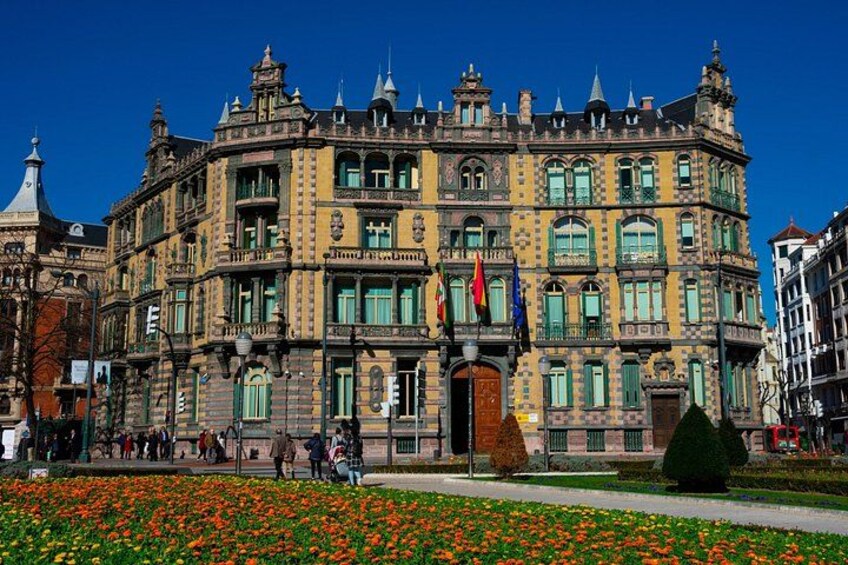 Un edificio histórico con arquitectura ornamentada, un jardín de flores en la fachada y banderas. 2