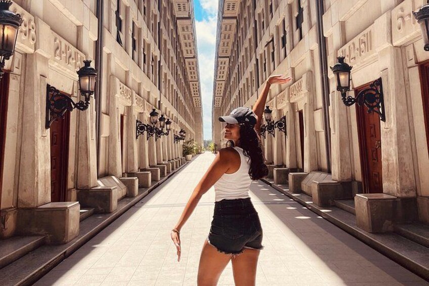 A person walking down a wide, sunlit street flanked by tall buildings with ornate street lamps. 3