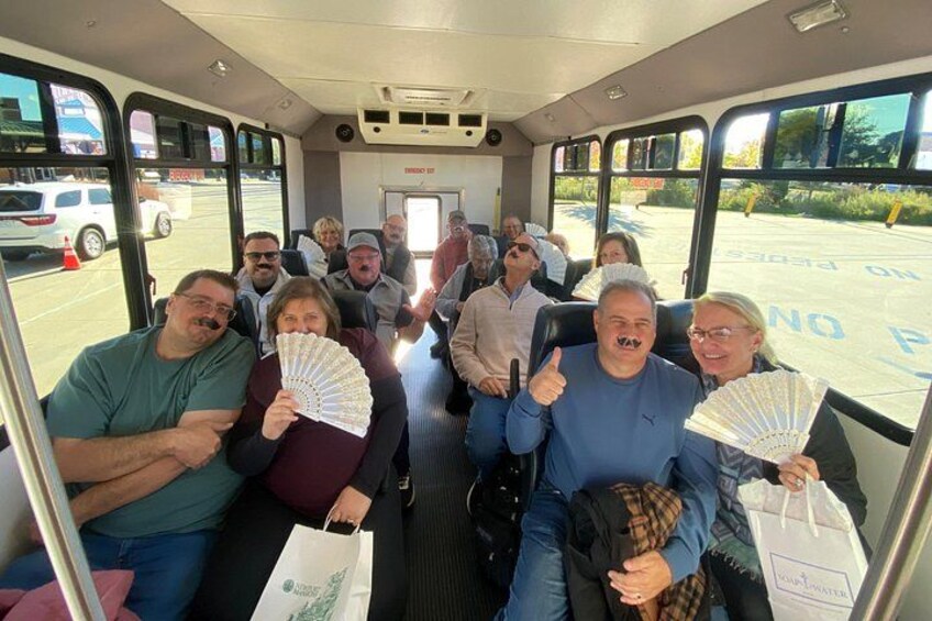 A group of people seated inside a bus, holding fans, with a parking lot visible through the windows. 3