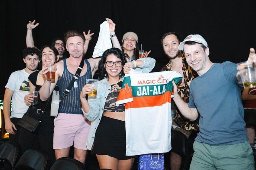 A group of people at an event, some holding drinks and one wearing a 'Magic City JAI-ALA' shirt. 2