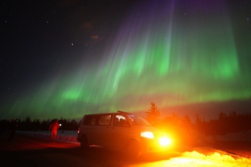 Ein Fahrzeug mit eingeschalteten Scheinwerfern, eine Person steht daneben, und im Hintergrund sind die Nordlichter zu sehen. 3