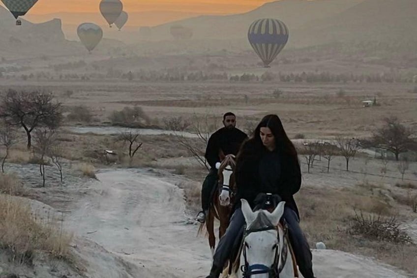 A dirt road with people riding horses, surrounded by open fields and distant hot air balloons. 2