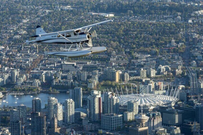 A seaplane flying over a city with a large stadium and water body below. 3