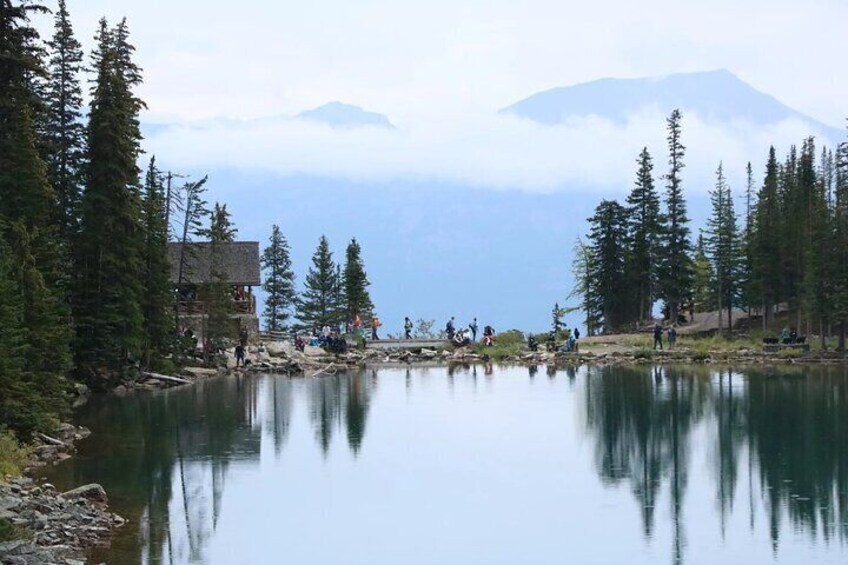 A calm lake surrounded by evergreen trees, with a mountain in the background and a small building near the water's edge. 5