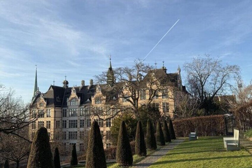 Un grand bâtiment historique, doté d’une tour et d’une flèche, entouré d’un jardin bien entretenu, avec des haies taillées et une pelouse. 5
