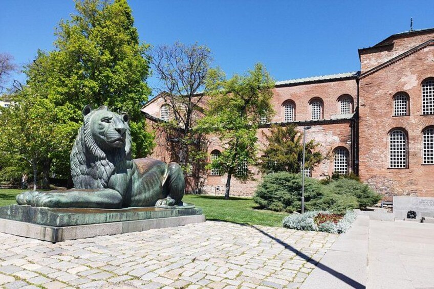 A bronze lion statue in a park with a brick building in the background. 3