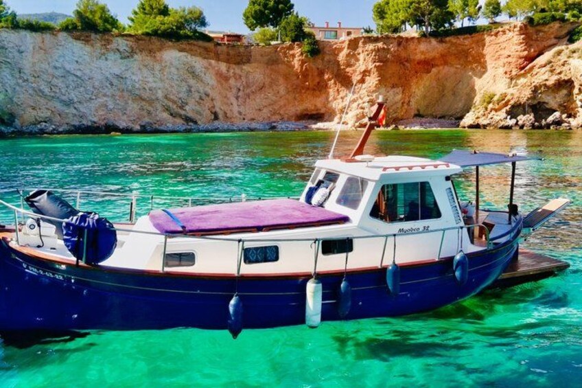 Een boot genaamd 'Moabro 38' ligt aangemeerd aan een heldere, turquoise oever, met een rotsachtige klif op de achtergrond. 1