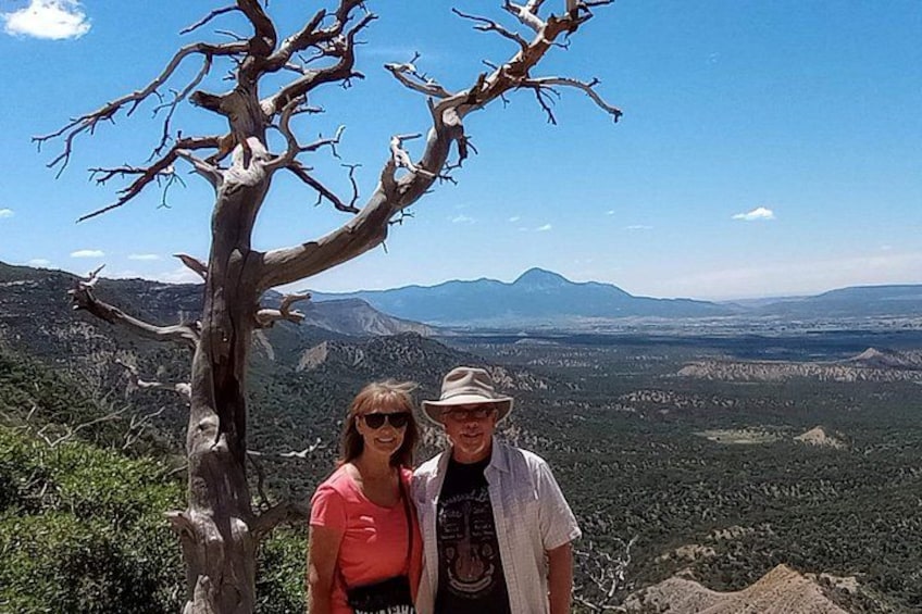 A man and a woman standing near a dead tree with a mountainous landscape in the background. 2
