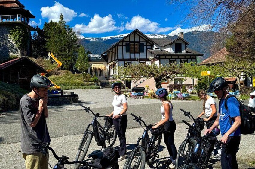 A group of people with bicycles in a town square, a mountain in the background, and traditional buildings. 2