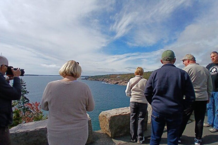 En grupp människor står vid en klippig strand och blickar ut över en vattenyta och ett avlägset landområde. 4