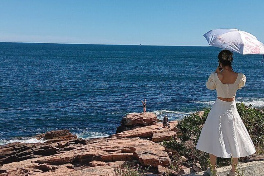 En kvinna i en vit klänning står på en stenig strand och håller ett parasoll, med utsikt över havet. 1