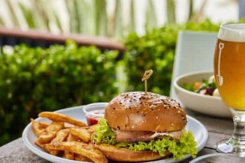 Hamburguesa con pan de sésamo, lechuga y un palillo, acompañada de papas fritas y una cerveza en una mesa. 2