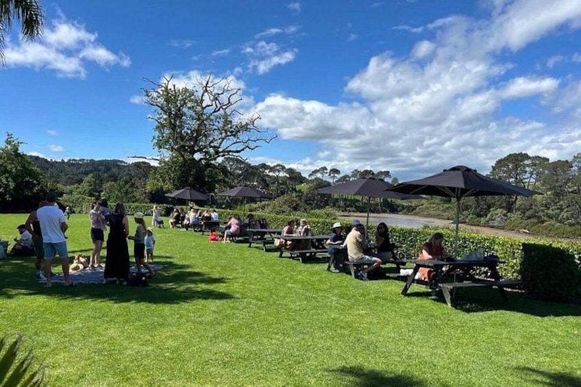 Un parque con mesas de picnic y sombrillas, gente sentada y caminando, y un gran árbol al fondo. 4
