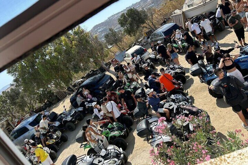 Eine Gruppe von Personen versammelt sich um Motorräder auf einem Parkplatz. Im Hintergrund sind Blumen und Bäume zu sehen. 5