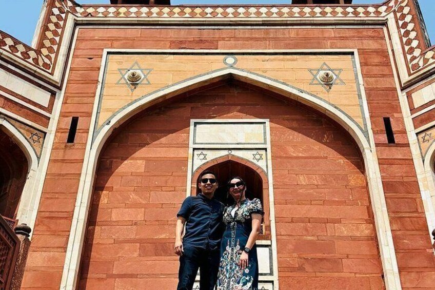 A man and a woman standing in front of a red brick building with a large archway and decorative elements. 2