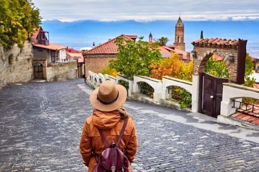 Una persona con cappello e giacca che cammina su una strada acciottolata, sullo sfondo edifici e una torre. 4