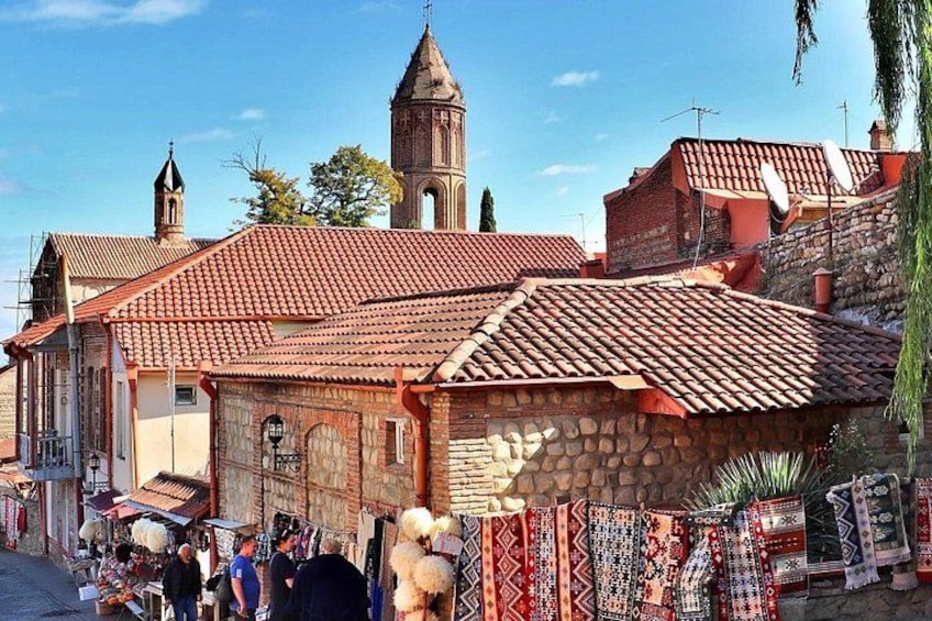 A street with traditional buildings, a church tower, and a market with patterned textiles. 1