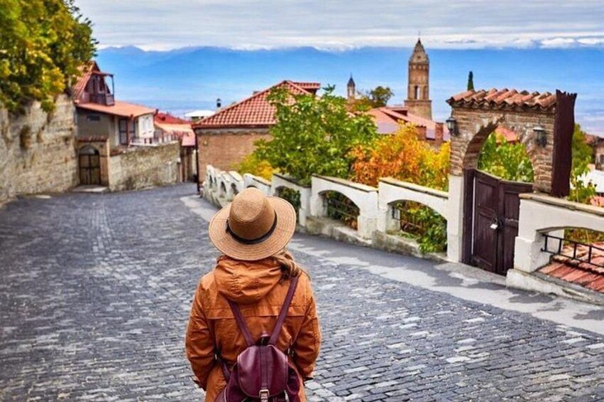 A person in a hat and jacket walking on a cobblestone street, with buildings and a tower in the background. 5
