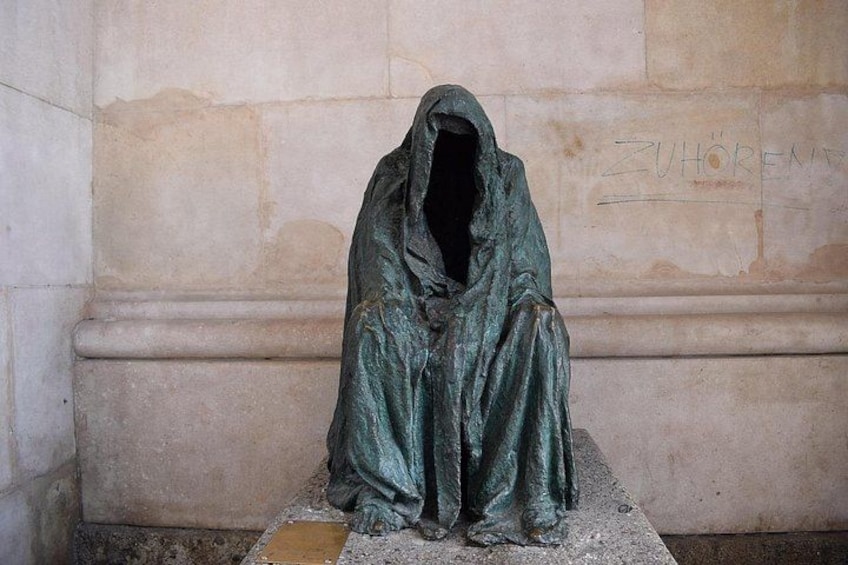 A statue of a hooded figure seated on a pedestal against a stone wall with the inscription 'ZUHLEREN'. 5