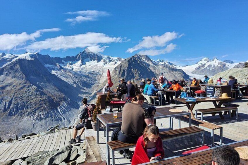 A group of people sitting at wooden tables on a wooden deck, overlooking a mountain range with snow-capped peaks. 1