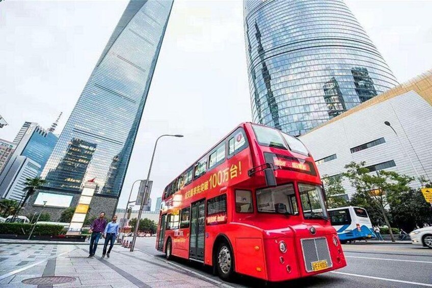 A red double-decker bus on a city street with modern skyscrapers in the background. 1