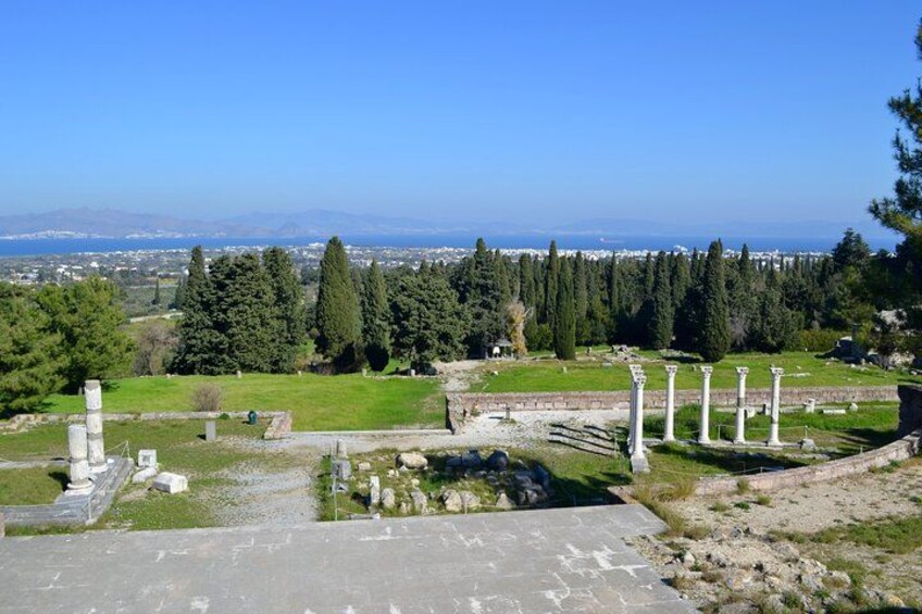 Ancient ruins with columns, a grassy area, and a view of the sea and city in the distance. 4