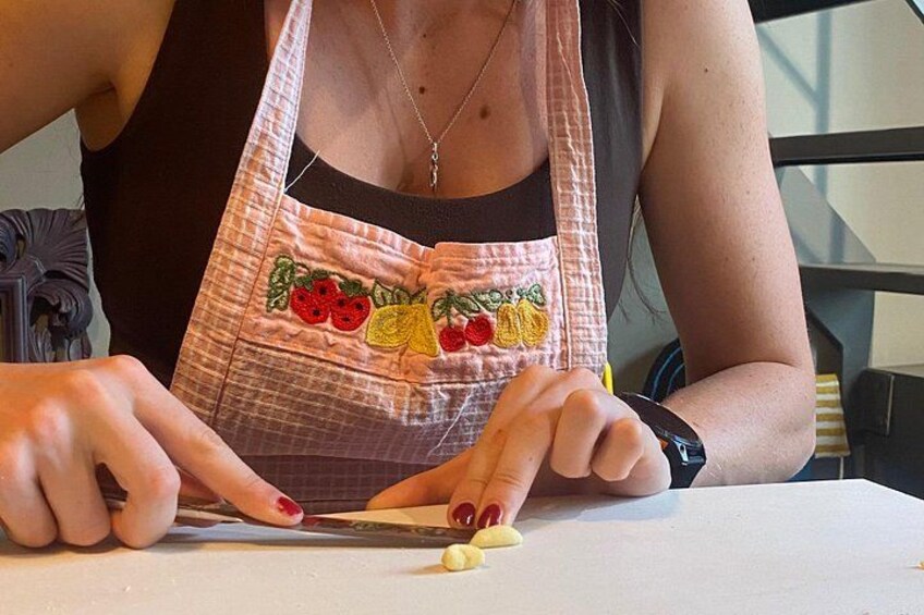 A person wearing a pink apron with fruit embroidery, holding a knife and cutting a yellow vegetable on a white table. 2